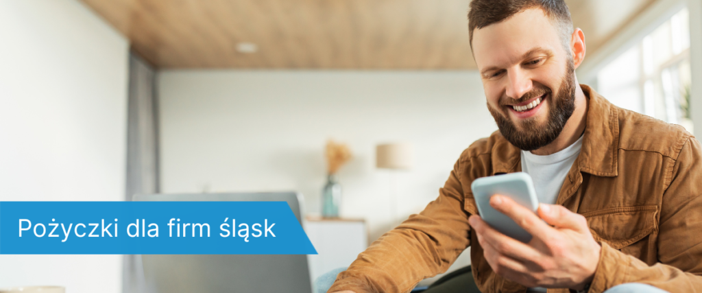 Mężczyzna z brodą w brązowej kurtce siedzi na kanapie z laptopem i telefonem, uśmiecha się, patrząc na ekran